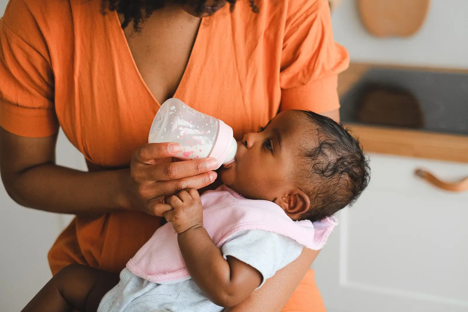 A woman in an orange shirt holding a feeding bottle for a baby