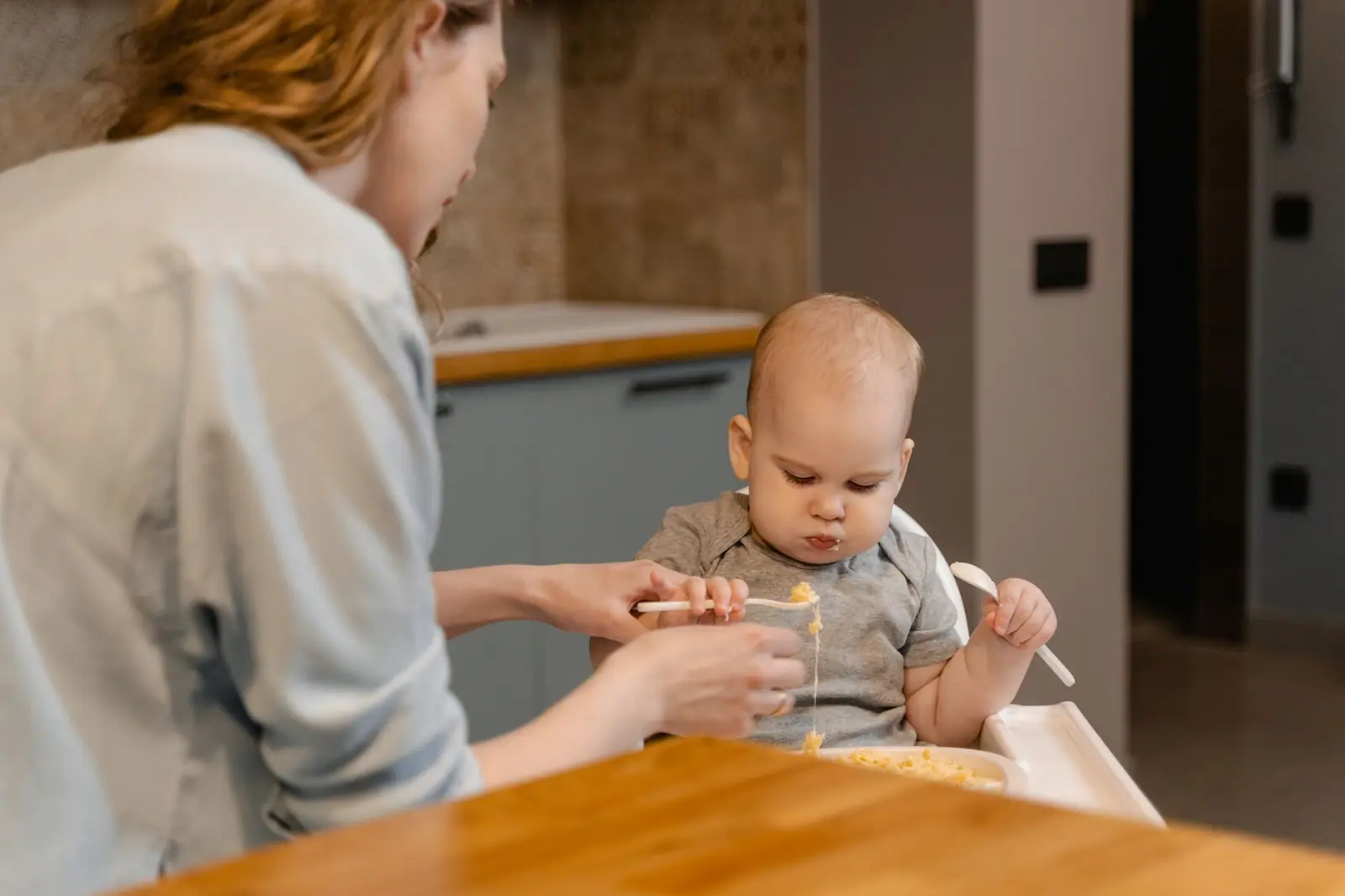 An adult in a gray top spoon-feeding a baby with a gray bib in a high chair