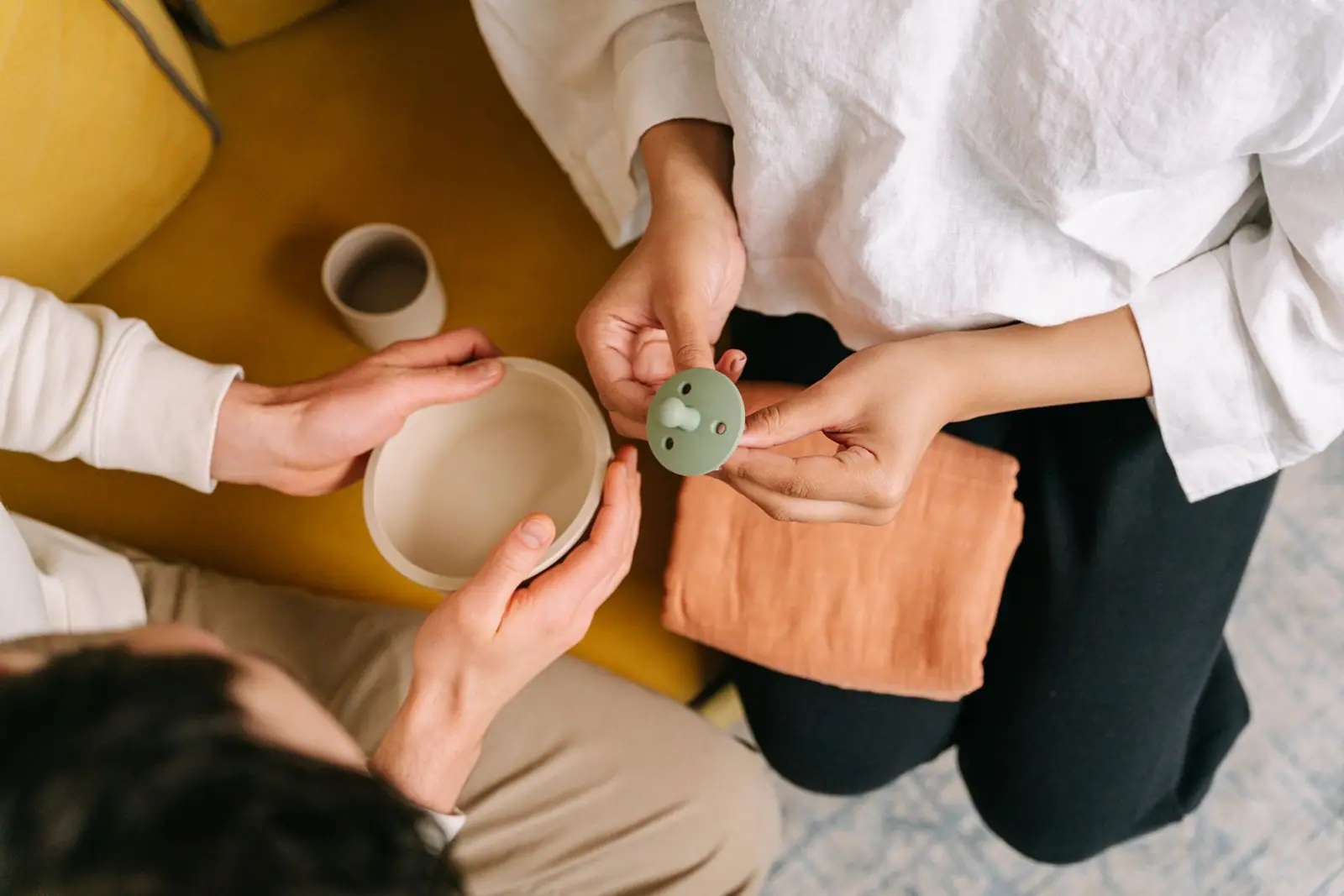 An adult holding a green pacifier near a baby and feeding supplies