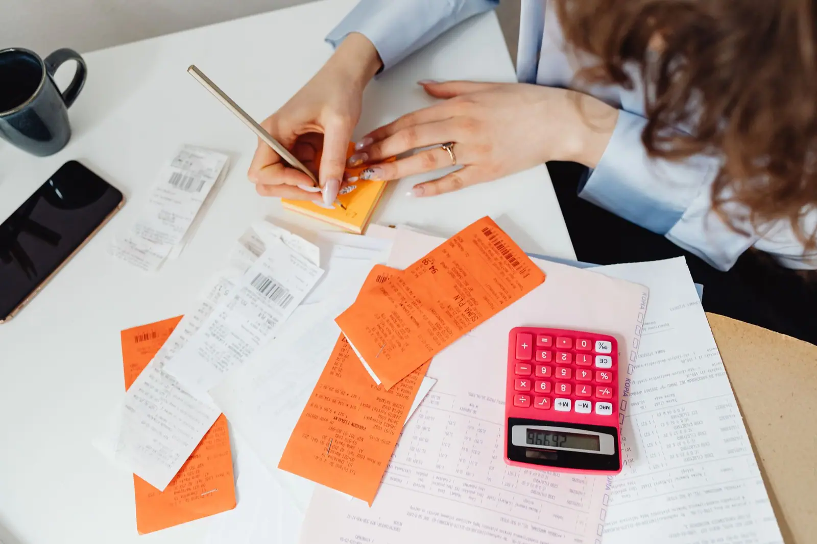 An adult writing on financial paperwork beside orange notes and a red calculator