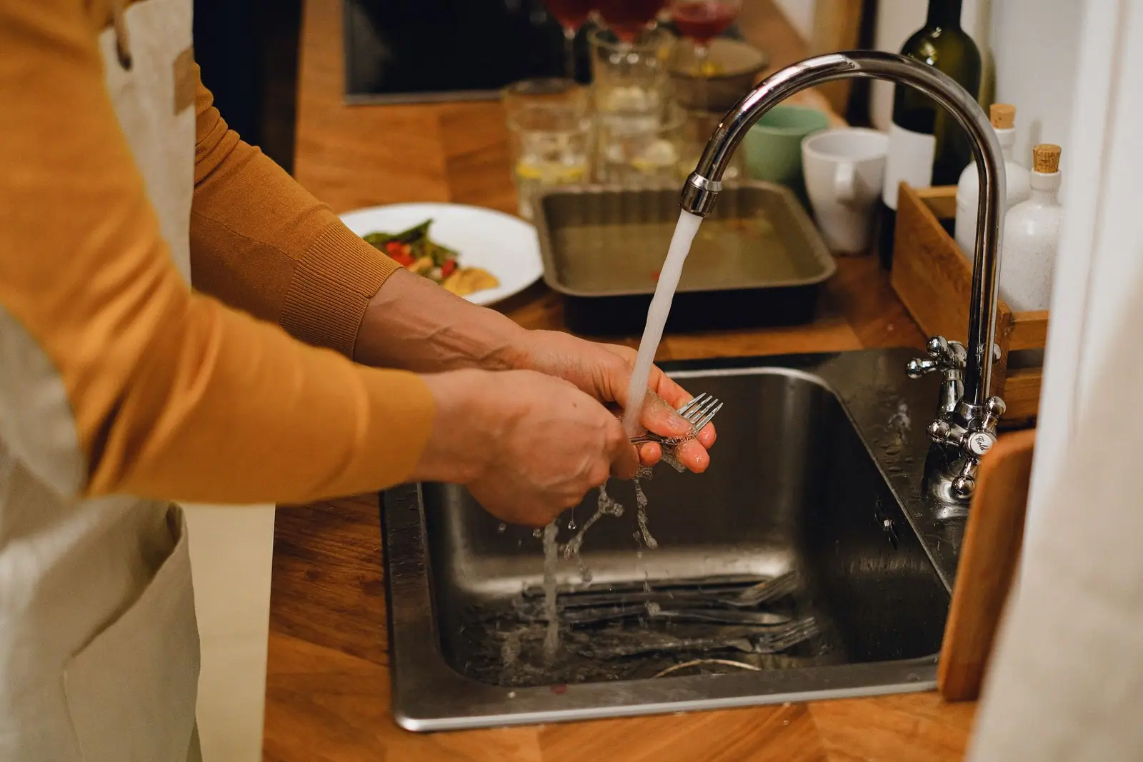An adult washing utensils under running water at a kitchen sink